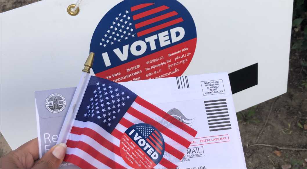 I Voted sticker on an American Flag, on top of a vote-by-mail ballot